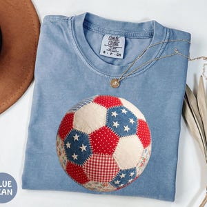 May include: Blue jean colored t-shirt with a patchwork soccer ball design in red, white, and blue. The shirt has a crew neck and short sleeves. A gold necklace with a pendant is draped on the shirt. The tag reads "Comfort Colors".