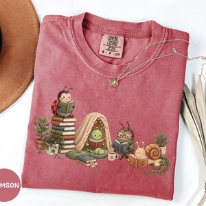 May include: A crimson short-sleeve t-shirt featuring a graphic of a ladybug, a caterpillar in a tent, a beetle with glasses, and a snail, all reading books. The shirt has a crew neck and a Comfort Colors label indicating size small.