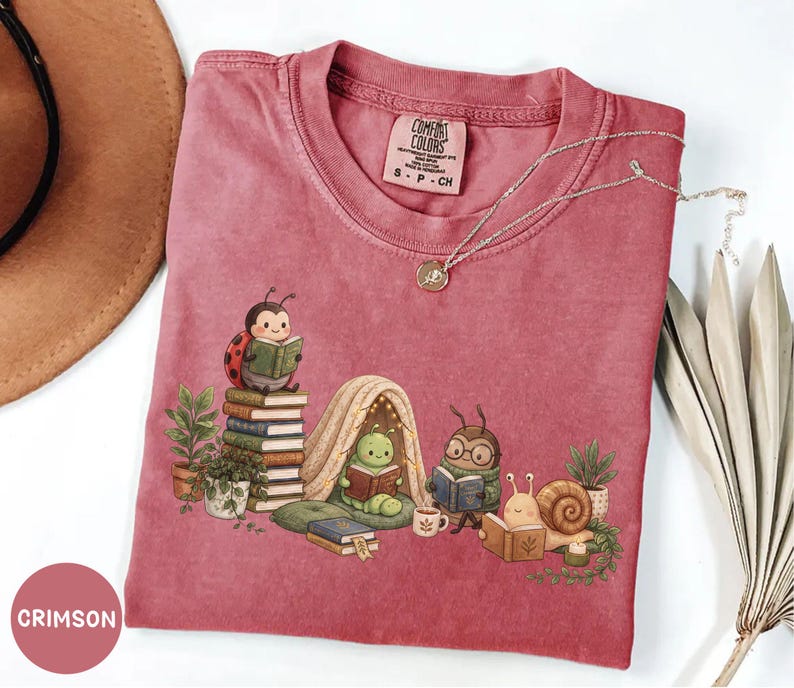 May include: A crimson short-sleeve t-shirt featuring a graphic of a ladybug, a caterpillar in a tent, a beetle with glasses, and a snail, all reading books. The shirt has a crew neck and a Comfort Colors label indicating size small.