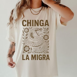 Puede incluir: Camiseta color marfil con una ilustraci&oacute;n de serpiente marr&oacute;n y el texto "CHINGA LA MIGRA". El dise&ntilde;o incorpora motivos florales. La camiseta es de corte holgado.
