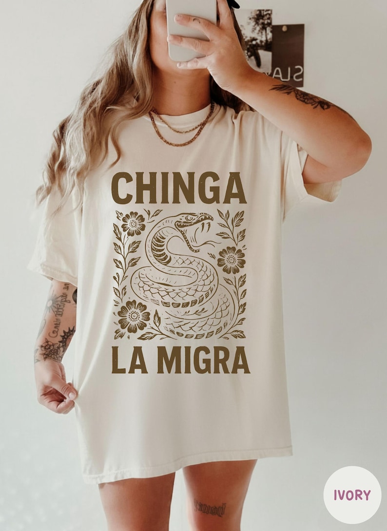 Puede incluir: Camiseta color marfil con una ilustraci&oacute;n de serpiente marr&oacute;n y el texto "CHINGA LA MIGRA". El dise&ntilde;o incorpora motivos florales. La camiseta es de corte holgado.