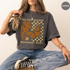May include: Dark gray t-shirt featuring a monarch butterfly design with orange and yellow accents. The text "THE ONLY ORANGE MONARCH I WANT" is printed above and below the butterfly. The shirt has short sleeves and a relaxed fit.