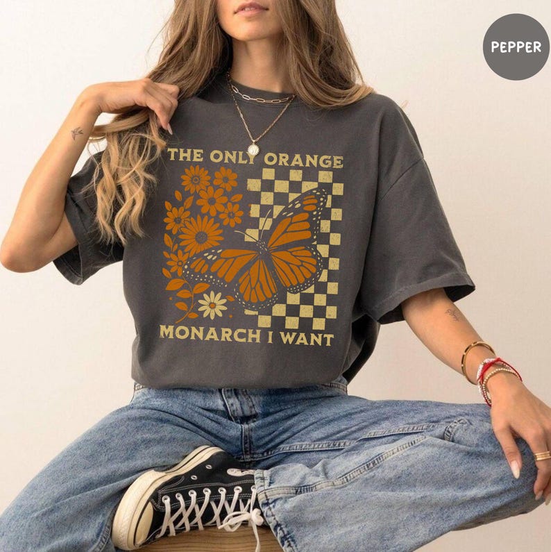 May include: Dark gray t-shirt featuring a monarch butterfly design with orange and yellow accents. The text "THE ONLY ORANGE MONARCH I WANT" is printed above and below the butterfly. The shirt has short sleeves and a relaxed fit.