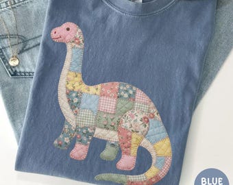 Puede incluir: Una camiseta azul con un gráfico de dinosaurio acolchado hecho de varios parches de tela floral y de cuadros vichy en tonos rosa, amarillo, verde y azul. La camiseta tiene cuello redondo con una etiqueta Comfort Colors y se combina con mezclilla azul.
