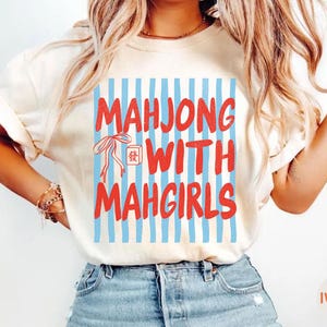 Peut inclure: Un t-shirt &agrave; manches courtes couleur ivoire avec un imprim&eacute; graphique de rayures verticales bleu clair. Le texte "MAHJONG WITH MAHGIRLS" est &eacute;crit en lettres rouges grasses, accompagn&eacute; d'un dessin au trait rouge d'un n&oelig;ud et d'une tuile.