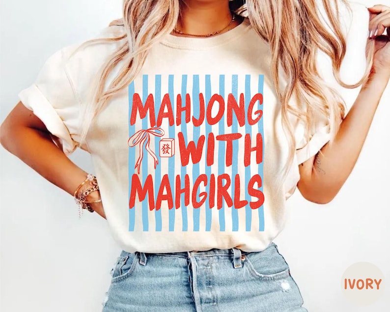 Peut inclure: Un t-shirt &agrave; manches courtes couleur ivoire avec un imprim&eacute; graphique de rayures verticales bleu clair. Le texte "MAHJONG WITH MAHGIRLS" est &eacute;crit en lettres rouges grasses, accompagn&eacute; d'un dessin au trait rouge d'un n&oelig;ud et d'une tuile.