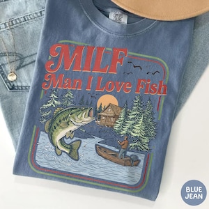 May include: A blue Comfort Colors t-shirt with a graphic that reads "MILF Man I Love Fish" in red and orange. The graphic features a fish jumping out of the water, a fisherman in a boat, and a cabin in the woods.