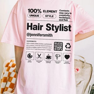 K&ouml;nnte beinhalten: Ein hellrosa T-Shirt mit schwarzem Text und Grafiken auf der R&uuml;ckseite. Das Design imitiert ein Produktetikett mit dem Text Hair Stylist, @jennifersmith, und einer Liste von Inhaltsstoffen wie kreativ, detailorientiert und Liebe zur Ver&auml;nderung.