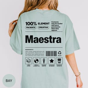 May include: A sage green t-shirt featuring a black graphic on the back that resembles a nutrition label. The text includes the word Maestra, a list of ingredients, and icons representing a book, globe, torch, star, and coffee cup with text labels.