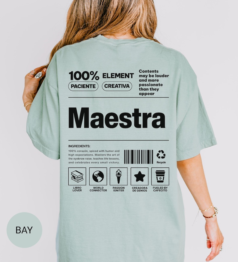 May include: A sage green t-shirt featuring a black graphic on the back that resembles a nutrition label. The text includes the word Maestra, a list of ingredients, and icons representing a book, globe, torch, star, and coffee cup with text labels.