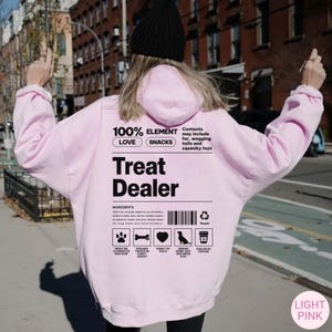May include: Light pink hoodie with the text "Treat Dealer" and ingredient list. The back of the hoodie has the text "100% Element Love Snacks" and icons of a dog, bone, heart, bird, and coffee cup. The hoodie is made of a soft material.