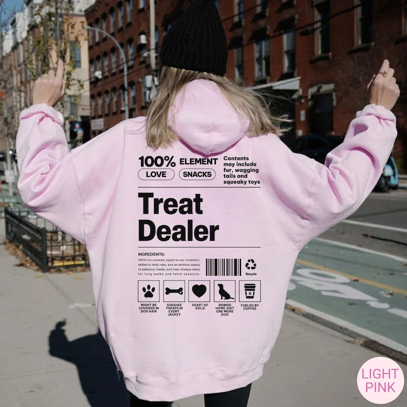 May include: Light pink hoodie with the text "Treat Dealer" and ingredient list. The back of the hoodie has the text "100% Element Love Snacks" and icons of a dog, bone, heart, bird, and coffee cup. The hoodie is made of a soft material.