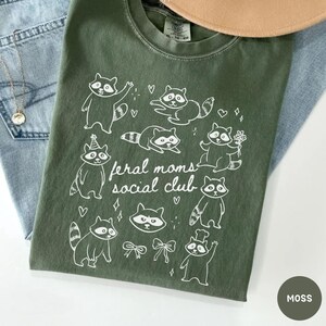 K&ouml;nnte beinhalten: Moosgr&uuml;nes T-Shirt mit wei&szlig;er Grafik von Waschb&auml;ren und dem Text "feral moms social club". Das Shirt ist auf einer blauen Jeans gefaltet. Eine goldene Halskette und ein beiger Hut sind ebenfalls sichtbar.