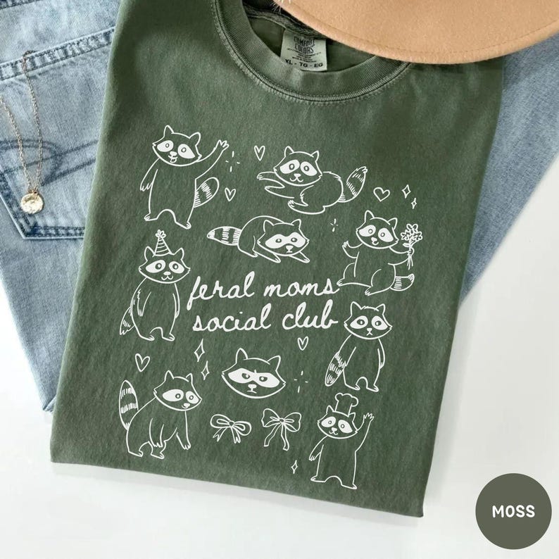 K&ouml;nnte beinhalten: Moosgr&uuml;nes T-Shirt mit wei&szlig;er Grafik von Waschb&auml;ren und dem Text "feral moms social club". Das Shirt ist auf einer blauen Jeans gefaltet. Eine goldene Halskette und ein beiger Hut sind ebenfalls sichtbar.
