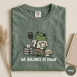 K&ouml;nnte beinhalten: Ein moosgr&uuml;nes T-Shirt mit einer gezeichneten Froschfigur, die eine Krawatte tr&auml;gt und einen Taschenrechner sowie ein Klemmbrett h&auml;lt. Das Motiv zeigt B&uuml;cherstapel mit der Aufschrift Tax, Invoices, Receipts, eine Tasse und den Text WE BALANCE AT DAWN.