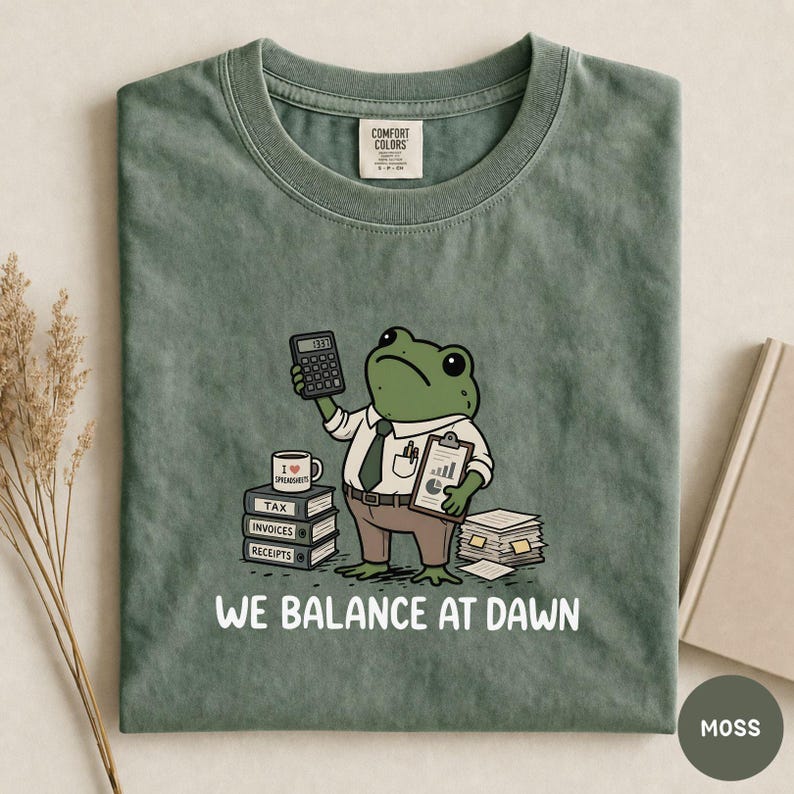 K&ouml;nnte beinhalten: Ein moosgr&uuml;nes T-Shirt mit einer gezeichneten Froschfigur, die eine Krawatte tr&auml;gt und einen Taschenrechner sowie ein Klemmbrett h&auml;lt. Das Motiv zeigt B&uuml;cherstapel mit der Aufschrift Tax, Invoices, Receipts, eine Tasse und den Text WE BALANCE AT DAWN.