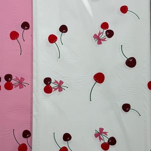 May include: White wrapping paper with a repeating pattern of red cherries with green stems and pink bows. The paper is partially covered with a clear plastic sheet.