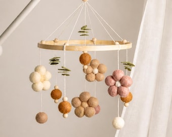 May include: A baby mobile featuring a wooden ring with cream, brown, and pink felt flowers and spheres. The mobile includes green felt leaf accents and is suspended from a white arm.