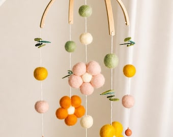May include: A baby mobile featuring felt flowers and spheres in yellow, pink, green, and white, suspended from a wooden arch. The mobile hangs from a white arm, with a natural wood finish.