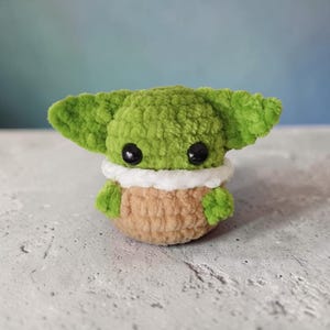 Peut inclure: Une petite peluche crochetée à la main avec une tête verte, de grandes oreilles pointues et des yeux en plastique noir. Le corps est de couleur beige avec une bordure blanche et duveteuse autour du cou, donnant un aspect doux et texturé.