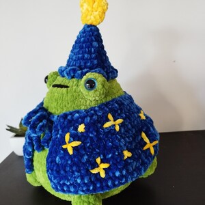 May include: A handmade green frog plushie wearing a blue wizard hat with a yellow star and a blue cape with yellow star accents. The frog has large, round eyes and is sitting on a black surface.