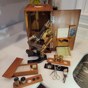 May include: An antique microscope with brass and black components, presented with its wooden storage case and various accessories. The case is open, revealing the microscope and its components. Additional items include small vials, scissors, and keys.