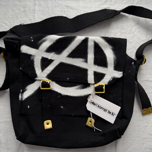 May include: Black messenger bag with a white anarchy symbol spray-painted on the flap. The bag has a black adjustable strap, gold-colored buckles, and a tag that reads "Don't interrupt the DJ-".