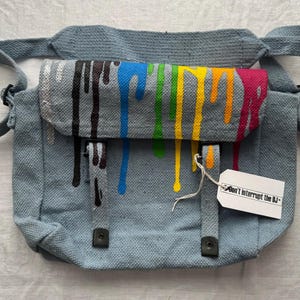 May include: A gray canvas messenger bag with a flap closure. The flap is decorated with colorful paint drips in black, blue, green, yellow, and pink. The bag has an adjustable strap and a tag that reads "Don't Interrupt the DJ".