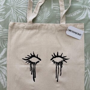 May include: Beige canvas tote bag featuring a black illustration of crying eyes. The bag has two beige handles and a tag that reads "Don't Interrupt the DJ". The tote bag is on a green and white patterned surface.