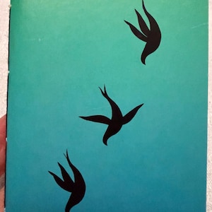 May include: A small notebook with a teal and blue gradient cover. The cover features three black bird silhouettes in flight. The notebook is held by a hand, showing the binding on the left side.