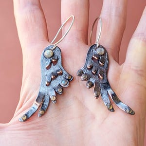May include: A pair of silver earrings with a unique design featuring a copper-toned, abstract wing shape with a small, iridescent gemstone at the top of each wing.