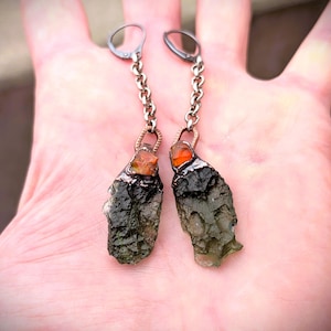 May include: A pair of dangle earrings featuring rough-cut, dark green and orange gemstones. The stones are suspended from silver-toned chains and lever-back ear wires. The earrings have a natural, organic aesthetic.