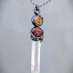 May include: A handcrafted pendant necklace featuring a clear quartz crystal point, accented with a yellow and red gemstone. The gemstones are encased in a twisted, dark metal setting, suspended from a silver chain.