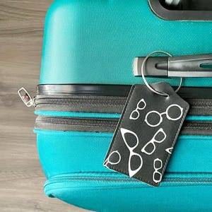 May include: Teal suitcase with a black zipper and handle. A black luggage tag with a white glasses pattern hangs from the handle. The tag is attached with a silver metal ring. The suitcase is on a wooden surface.