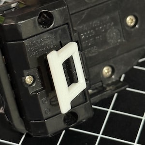 Puede incluir: Primer plano de un objeto de plástico negro con un clip blanco rectangular de extremo abierto adjunto. El clip está en primer plano y el objeto tiene tornillos y texto. El fondo es una cuadrícula en blanco y negro.