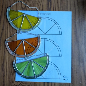 May include: Three stained glass citrus slice ornaments in yellow, orange, and green, each with a silver chain for hanging. The ornaments are on a wooden surface with a blue paper that says "Citrus Delight".