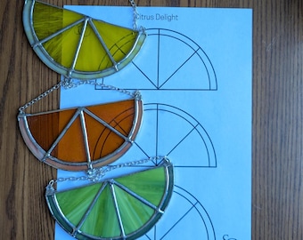 May include: Three stained glass citrus slice ornaments in yellow, orange, and green, each with a silver chain for hanging. The ornaments are on a wooden surface with a blue paper that says "Citrus Delight".