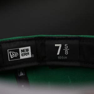 May include: Close-up of the interior of a green baseball cap. The black label displays the New Era logo and the size 7 5/8, equivalent to 60.6cm. The cap's interior is green.