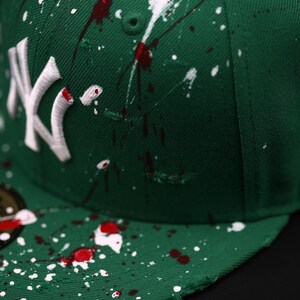 May include: Green baseball cap with white and red paint splatter design. Features the white embroidered "NY" logo. The cap has a curved brim and a button on top. The hat is a stylish accessory.