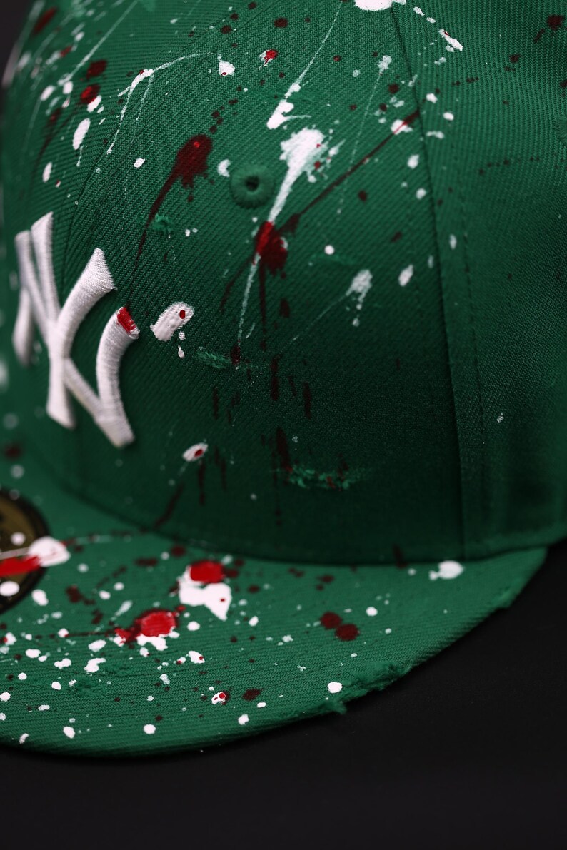 May include: Green baseball cap with white and red paint splatter design. Features the white embroidered "NY" logo. The cap has a curved brim and a button on top. The hat is a stylish accessory.