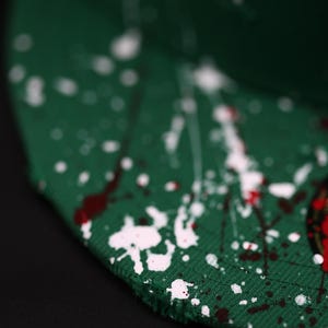 May include: Green baseball cap with white and red paint splatter design. The cap is made of a textured fabric and has a curved brim. The paint splatter adds a unique, artistic touch to the hat.