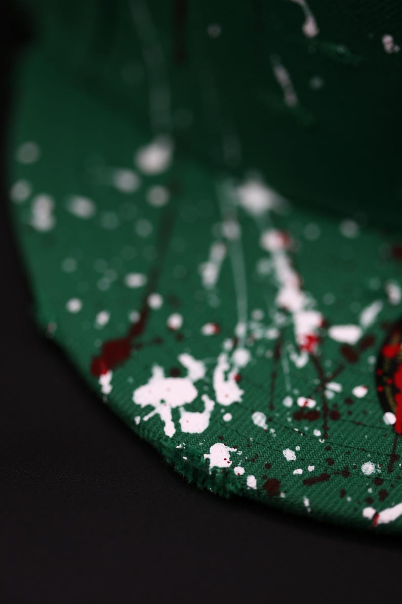May include: Green baseball cap with white and red paint splatter design. The cap is made of a textured fabric and has a curved brim. The paint splatter adds a unique, artistic touch to the hat.