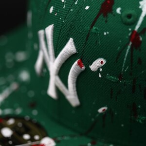 May include: Green baseball cap featuring a white embroidered "NY" logo and a paint splatter design in white and red. The cap has a curved peak. The style is reminiscent of streetwear fashion.
