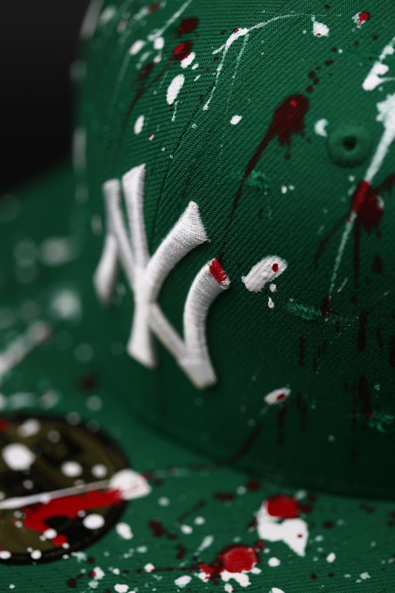 May include: Green baseball cap featuring a white embroidered "NY" logo and a paint splatter design in white and red. The cap has a curved peak. The style is reminiscent of streetwear fashion.