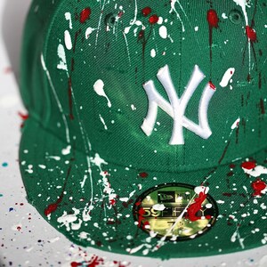 May include: A green baseball cap with white and red paint splatters. The cap features a white embroidered "NY" logo and a black and gold sticker on the brim. The hat is a fitted style.