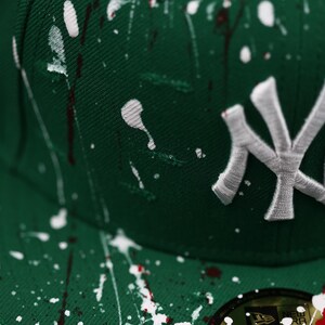 May include: A green baseball cap with white and red paint splatter. The cap features the New York Yankees logo in white embroidery. The brim has a sticker with the brand logo and size.