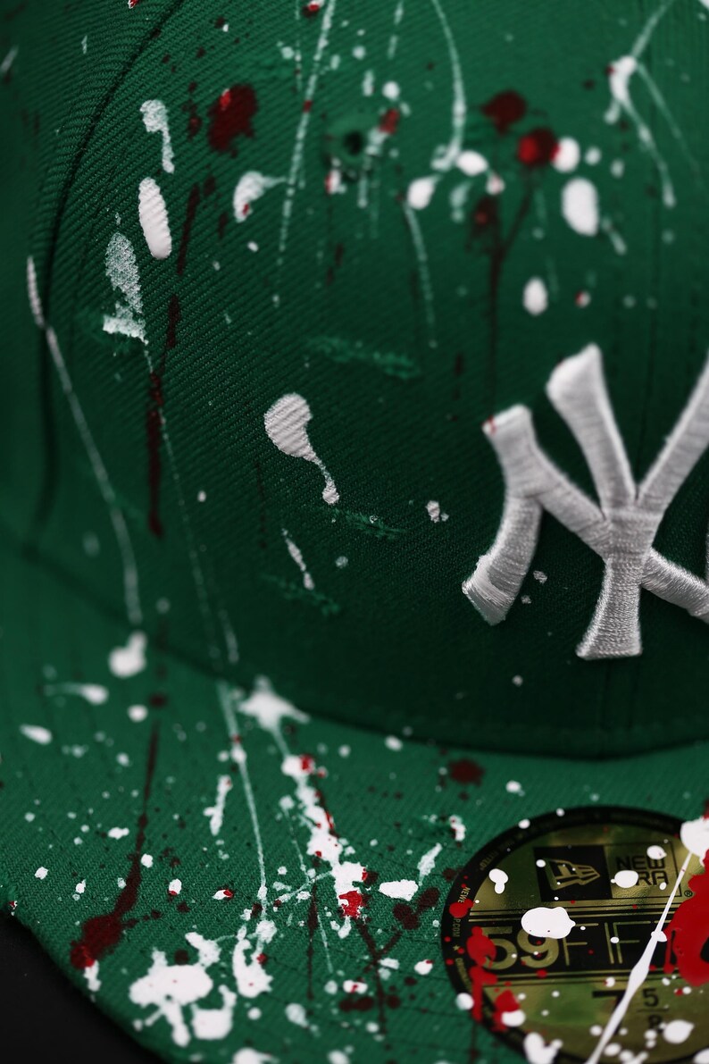 May include: A green baseball cap with white and red paint splatter. The cap features the New York Yankees logo in white embroidery. The brim has a sticker with the brand logo and size.