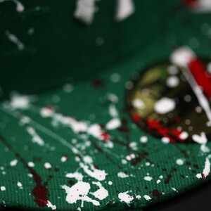 May include: Green baseball cap with white and red paint splatter design. The cap features a white embroidered logo. The brim has a distressed edge. The cap is a stylish accessory.