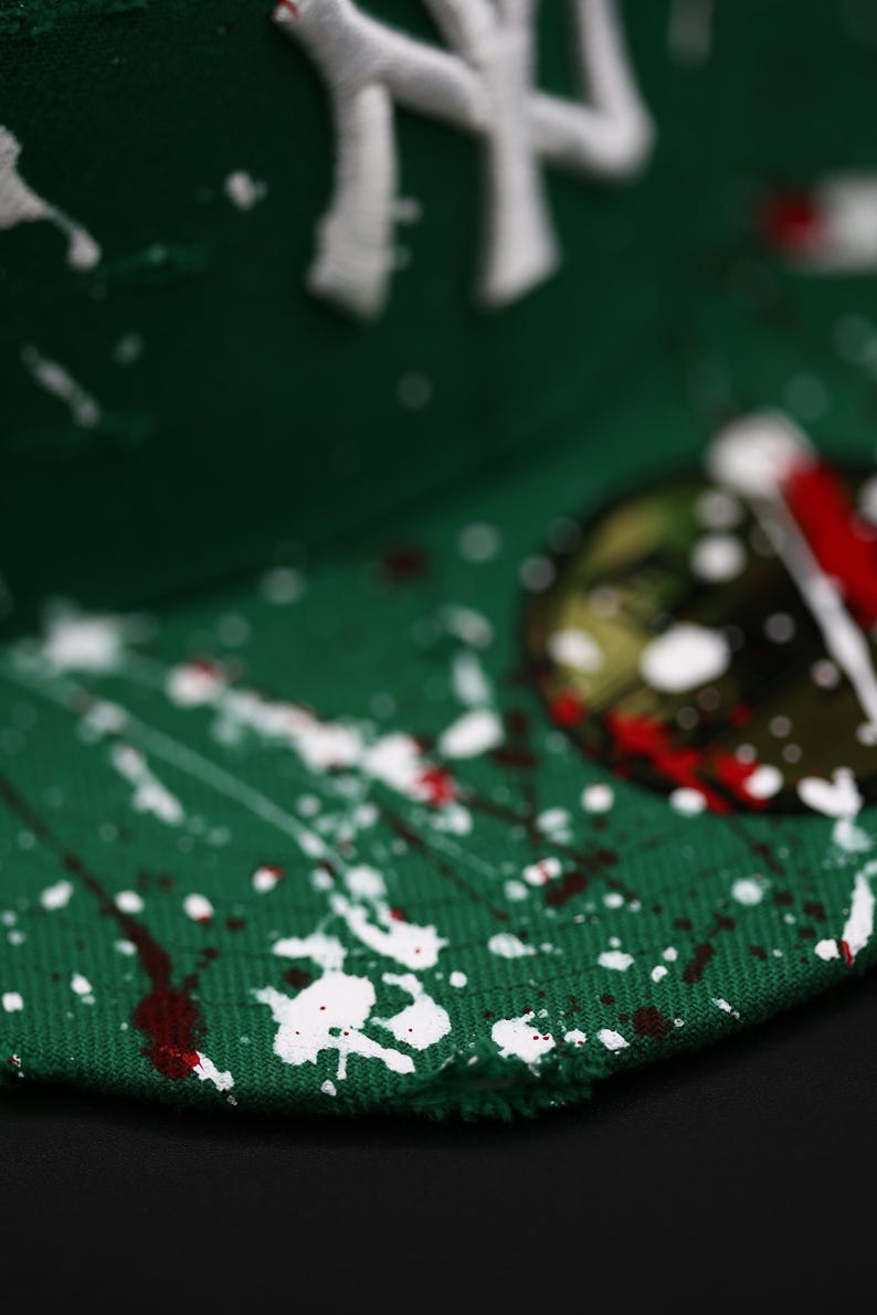 May include: Green baseball cap with white and red paint splatter design. The cap features a white embroidered logo. The brim has a distressed edge. The cap is a stylish accessory.