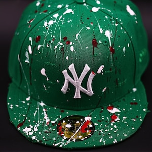 Op de afbeelding: Groene baseballcap met wit en rood verfspatterontwerp. Voorzien van een wit geborduurd "NY" logo op de voorkant. De cap heeft een platte klep en een aansluitend ontwerp, met een kleine sticker op de klep.